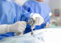 Surgeons hands close up performing surgery. Surgeon using drill machine during procedure. Doctors are wearing protective workwear in operating room.